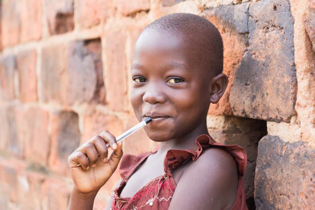 An 11-year-old Ugandan girl smiling, holding a pen against her mouth and leaning against a brick wall looking at the camera